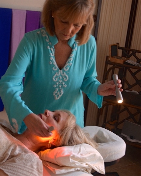 Lady wearing a turquoise shirt holding lights up to a lady's cheek while laying down.