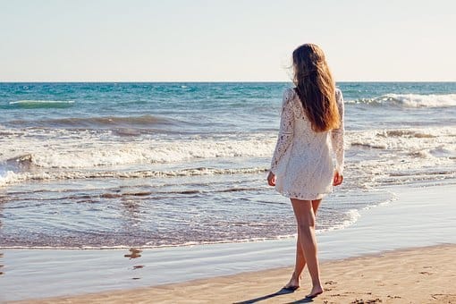 A lady on the beach wearing a lacey dress next to blue ocean with foamy waves.