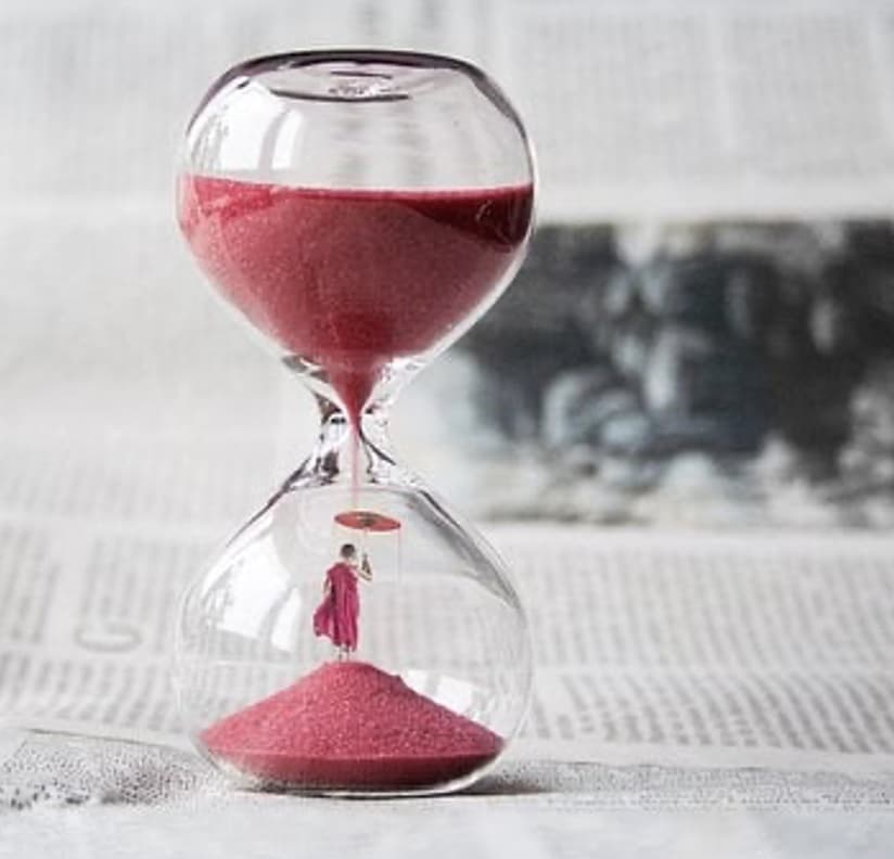 An hour glass with red sand pouring down and a newspaper behind it. There is person holding an umbrella under the sand in the bottom.